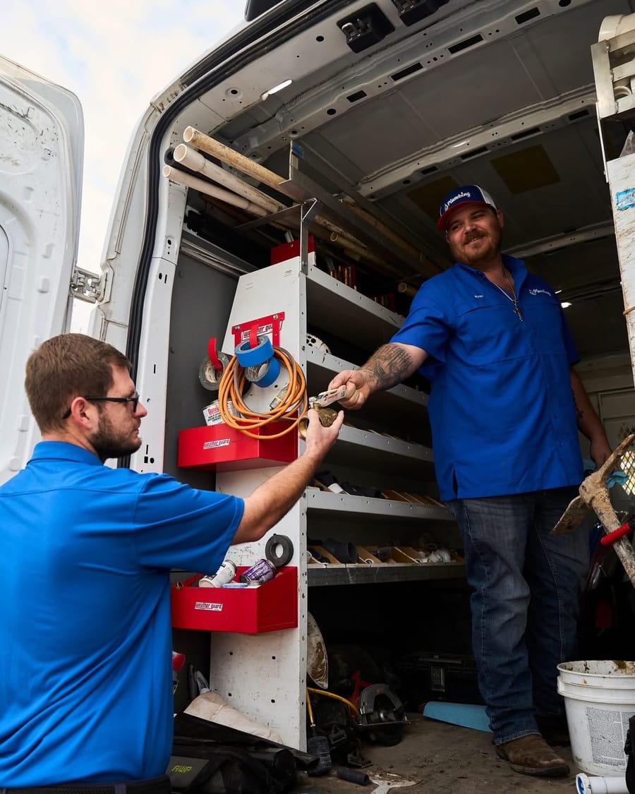 A plumber in a blue uniform hands a tool to a colleague from the back of a well-stocked service van.