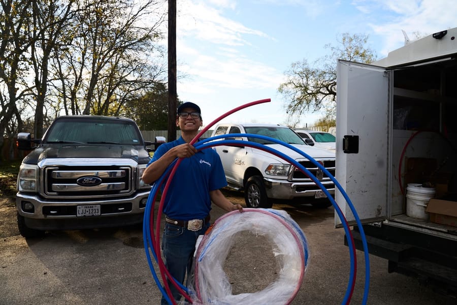 A smiling plumber in Seguin holding blue and red piping in front of service vehicles and a work truck.
