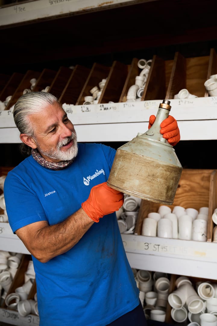 A plumber in a blue shirt and orange gloves examines a plumbing part in a workshop with shelves storing various pipe fitting.
