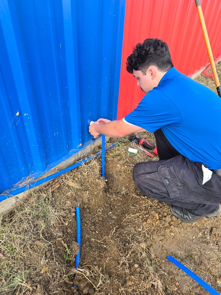 A plumber in a blue shirt is working outside on a blue pipe near a red and blue metal wall, surrounded by soil and tools, in Seguin.