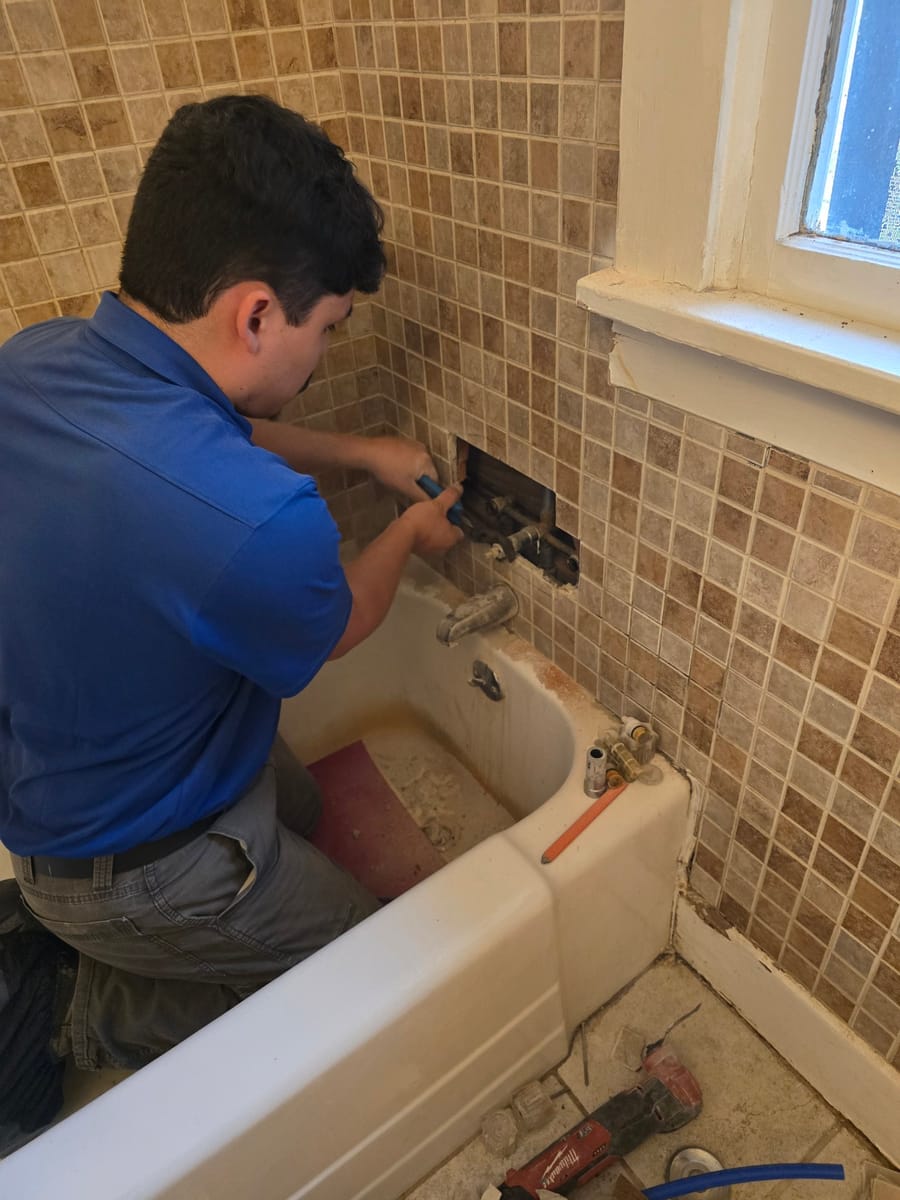 Plumber in a blue shirt repairing bathroom pipes behind a tiled wall.