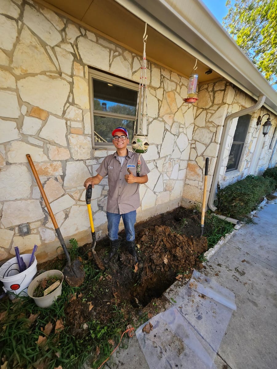 A plumber working outside a stone house in Seguin, standing next to a dug trench with tools and equipment.