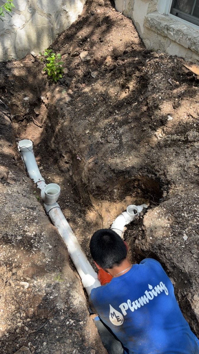 A plumber wearing a blue uniform with a plumbing company logo works on installing white PVC pipes in a dug trench next to a building.