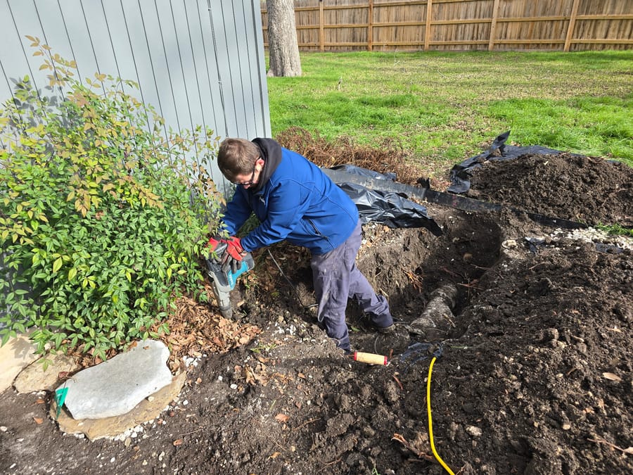 A plumber in a blue jacket and red gloves works with a power tool on an outdoor plumbing project.