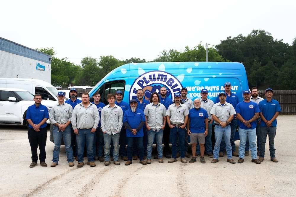 Group of water‑heater technicians in blue and gray shirts posing in a parking lot in front of a blue plumbing van with a company logo.